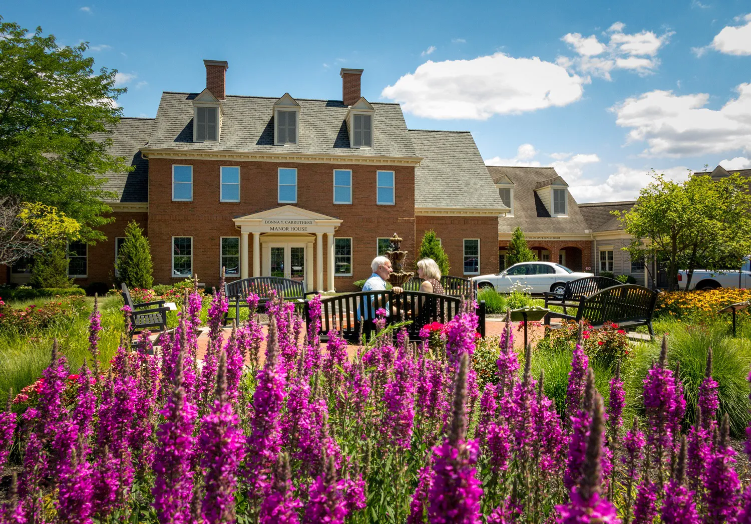 Exterior view of Manor House with older adult male and female sitting on a bench talking near the landscaped grounds